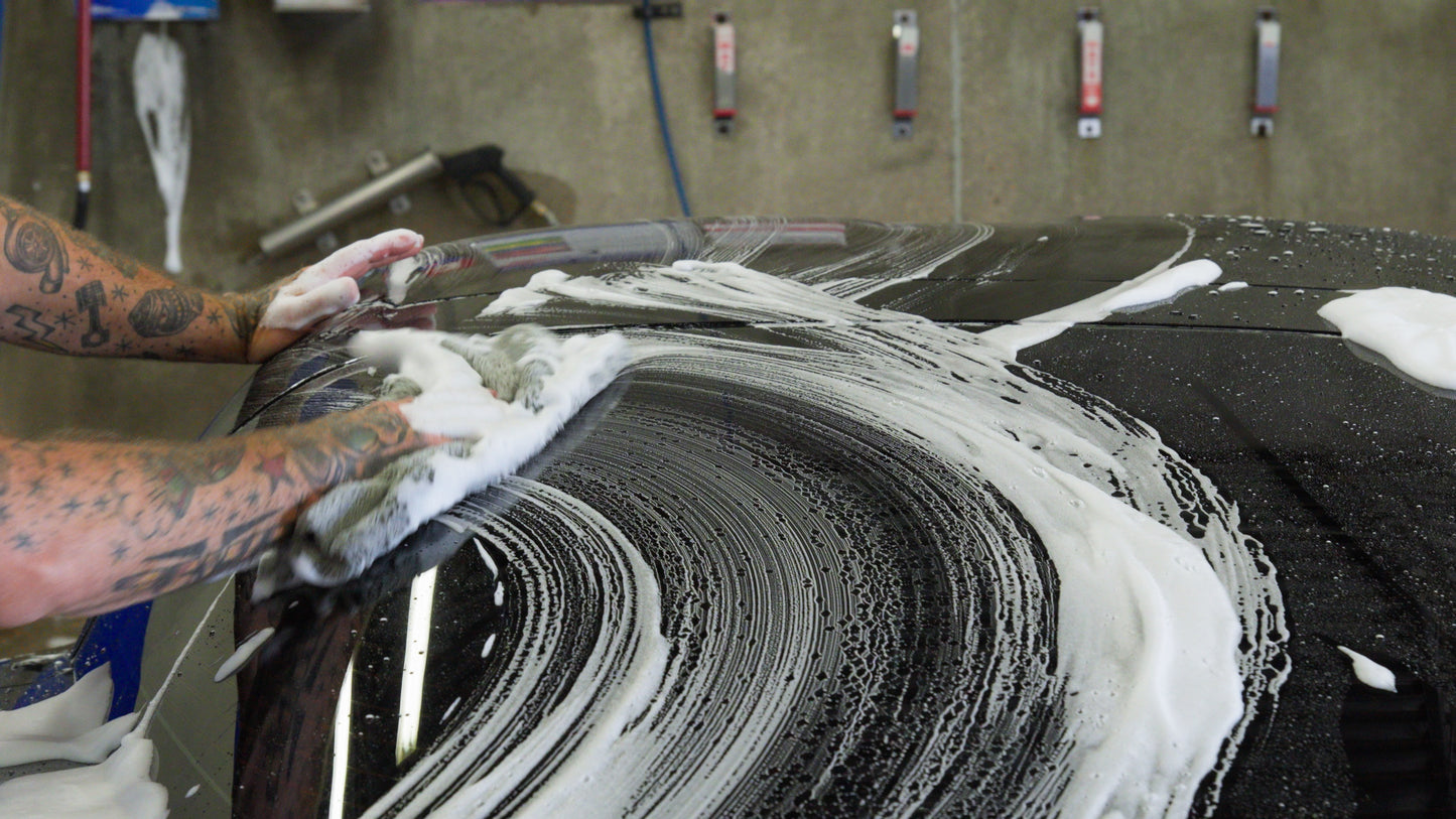 Person washing a car with soapy hands and arms, focusing on the soap suds on the car's surface.